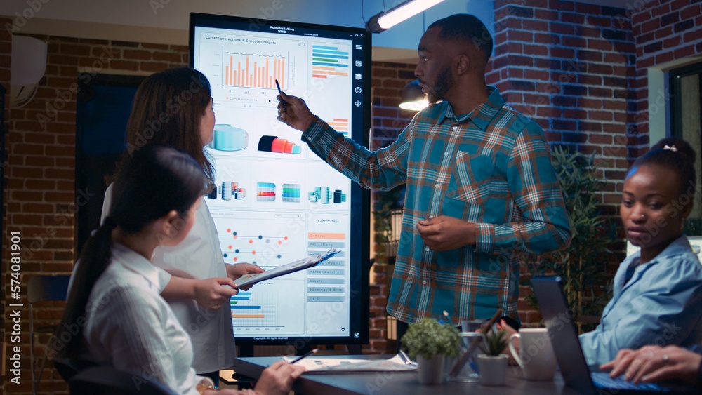 Diverse anaylsts presenting company marketing statistics visualization in business meeting, employees listening to presentation. Office workers in coworking space boardroom at night time