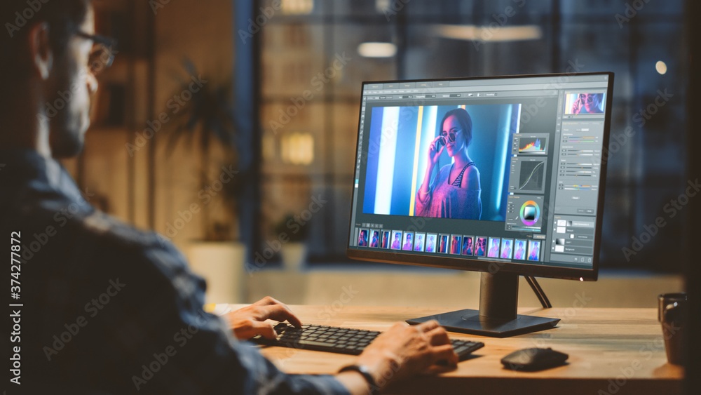 Over the Shoulder: Creative Young Digital Editor Works in Photo Editing Software on His Personal Computer with Big Display. In the Background Evening in Modern Loft Studio Office