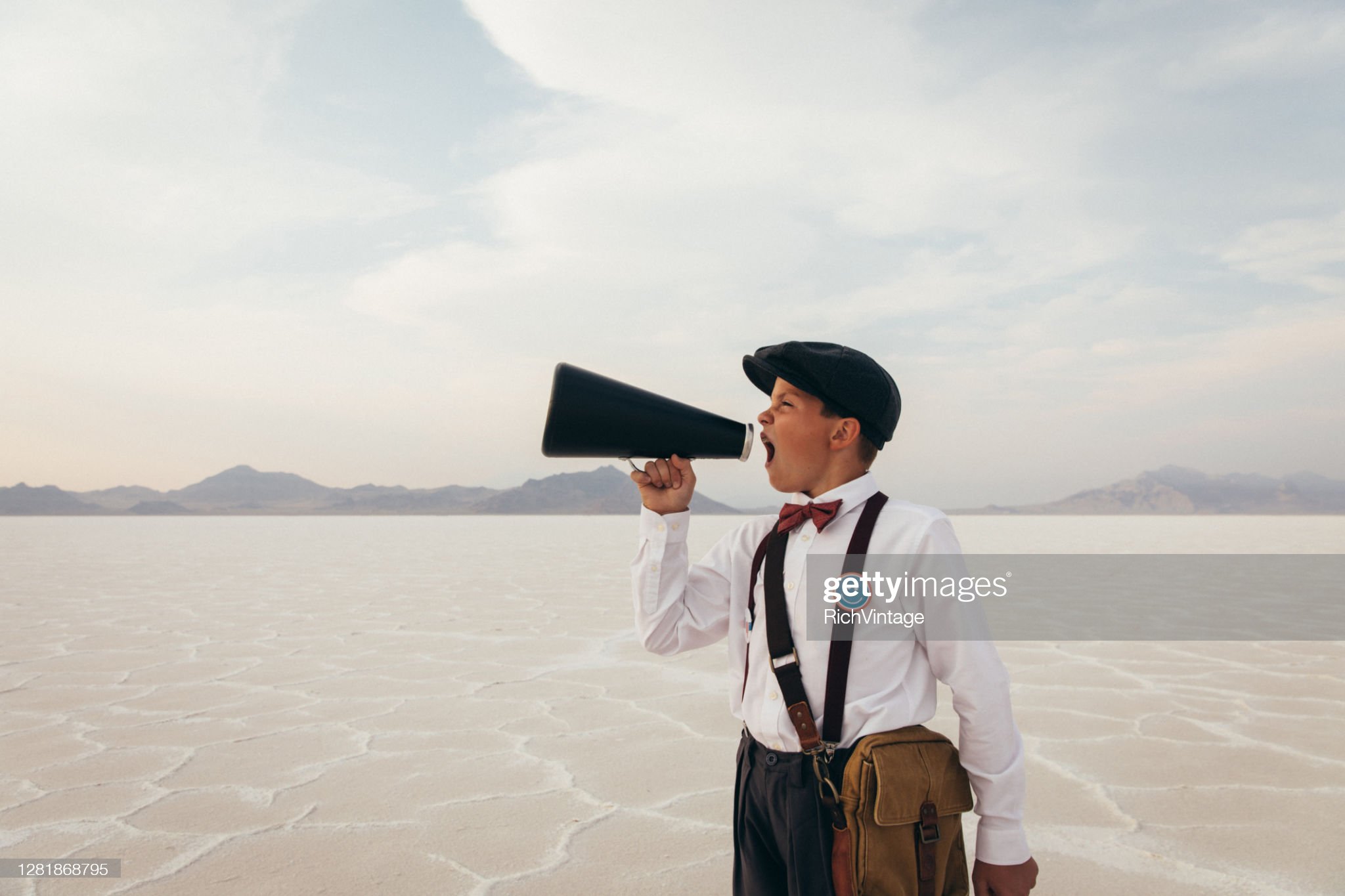 A young newspaper boy and teenager stands on the remote salt flats in Utah during the coronavirus pandemic trying to deliver his message. He is wearing old fashioned clothing with a face mask and a megaphone.